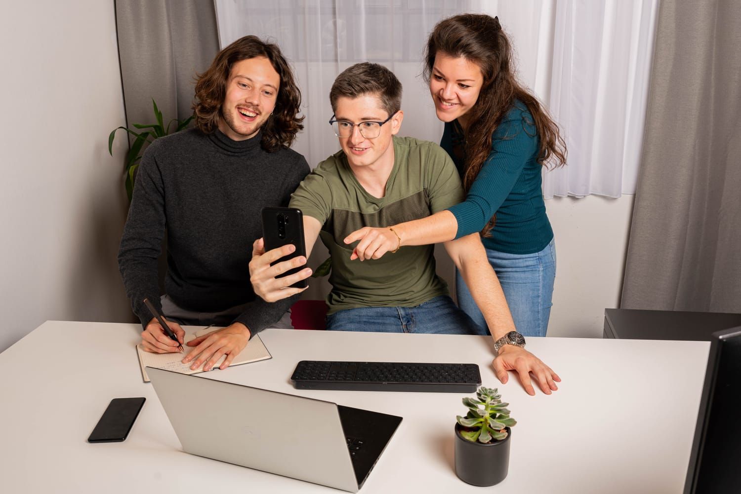 Teamfoto CodingBase, 3 Personen lachend auf ein Handy blickend und vor einem Laptop sitzend