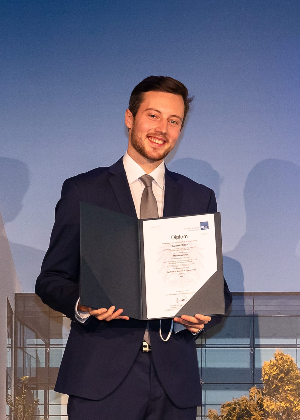 Ein Portrait unseres Geschäftsführers Raphael Posch, lächelnd mit Diplom in der Hand